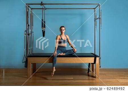 A young girl does Pilates exercises with a bed reformer, barrel machine tool. Beautiful slim fitness trainer on the background of a reformer doing various exercises gray, low key, light art. Fitness 62489492