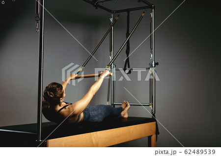 A young girl does Pilates exercises with a bed reformer, barrel machine tool. Beautiful slim fitness trainer on the background of a reformer doing various exercises gray, low key, light art. Fitness A young girl does Pilates exercises with a bed reformer, barrel machine tool. Beautiful slim fitness trainer on the background of a reformer doing various exercises gray, low key, light art. Fitness 62489539