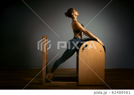 A young girl does Pilates exercises with a bed reformer, barrel machine tool. Beautiful slim fitness trainer on a background of a reformer gray, low key, light art. Fitness concept, healthy lifestyle. 62489561
