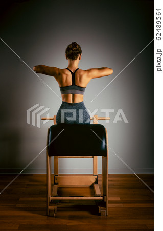 A young girl does Pilates exercises with a bed reformer, barrel machine tool. Beautiful slim fitness trainer on a background of a reformer gray, low key, light art. Fitness concept, healthy lifestyle. 62489564