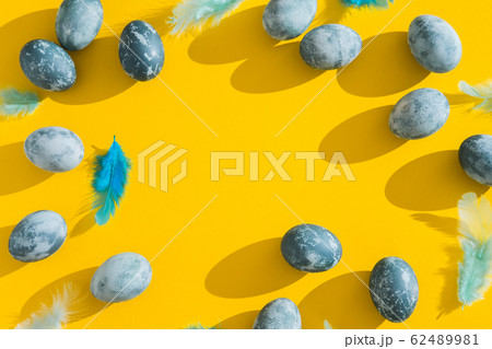 Blue textured colored Easter eggs and chicken bird feathers on a bright yellow holiday background backdrop. Spring holiday concept. Copyspace. Flat lay. Top view. Hard light. Long shadow. 62489981