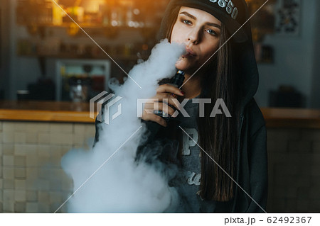 Young pretty woman in cap smoke an electronic cigarette at the vape shop. Hip-hop style. Closeup. 62492367