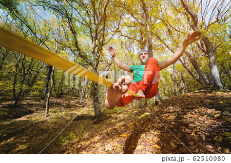 Wide angle male tightrope walker balancing while sitting barefoot on slackline in autumn forest. The concept of outdoor sports and active life of people aged 62510980