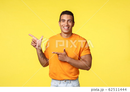 Cheerful modern man with tattooed arm, pointing left as promoting online store, invite guest join party, smiling joyfully, recommend try something, give advice what place visit, yellow background 62511444