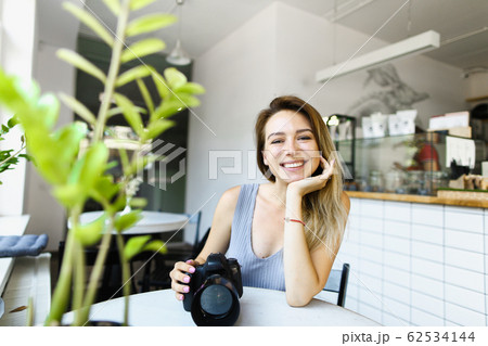 Young female smiling photographer sitting at cafe with camera and waiting for model. Young female smiling photographer sitting at cafe with camera and waiting for model. 62534144