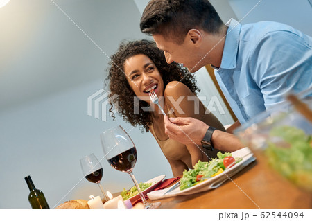 Romantic Date. Young multiethnic couple at home sitting at table eating dinner husband feeding wife with salad smiling playful Romantic Date. Young multiethnic couple at home sitting at table eating dinner husband feeding wife with salad smiling playful 62544094