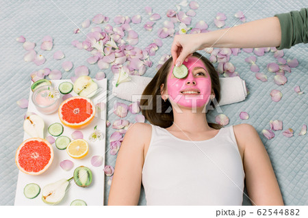 Beautiful young woman receiving facial mask of cucumber in beauty salon, hands of cosmetologist 62544882