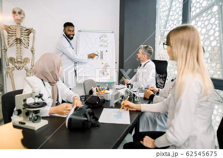 Cheerful Handsome young African doctor scientist smiling while standing near the classboard and showing new biochemical formulas for his multiracial colleagues at the meeting. Medicine, science 62545675