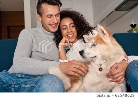 Romantic Date. Young multiethnic couple at home sitting on sofa playing with dog laughing cheerful 62548028