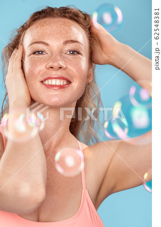 Summer Freestyle. Young woman with freckles standing isolated on blue soap bubbles touching hair posing cheerful 62548381