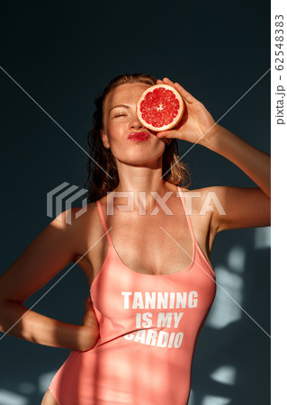 Summer Freestyle. Young woman with freckles and wet hair standing isolated on blue covering eye with grapefruit half posing grimace silly Summer Freestyle. Young woman with freckles and wet hair standing isolated on blue covering eye with grapefruit half posing grimace silly 62548383