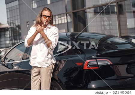 Mode of Transport. Man with long hair in sunglasses standing near electric car lighting cigarette looking aside cool Mode of Transport. Man with long hair in sunglasses standing near electric car lighting cigarette looking aside cool 62549034