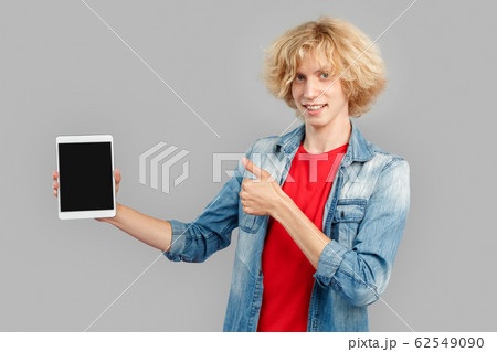 Freestyle. Teen blond boy standing isolated on gray showing screen on digital tablet copy space showing thumb up happy 62549090