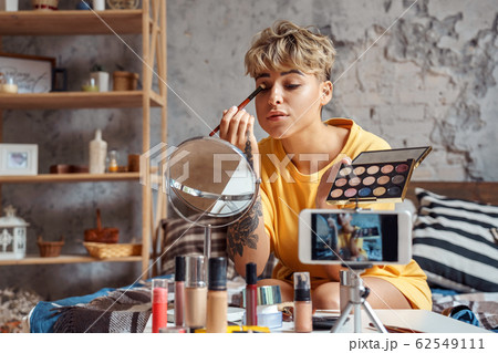 Make-up Blogger. Young woman sitting at stylish apartment recording video on smartphone looking at mirror applying eyeshadow concentrated 62549111