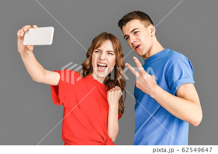 Freestyle. Young couple standing isolated on grey taking selfie on smartphone posing to camera gesturing playful Freestyle. Young couple standing isolated on grey taking selfie on smartphone posing to camera gesturing playful 62549647