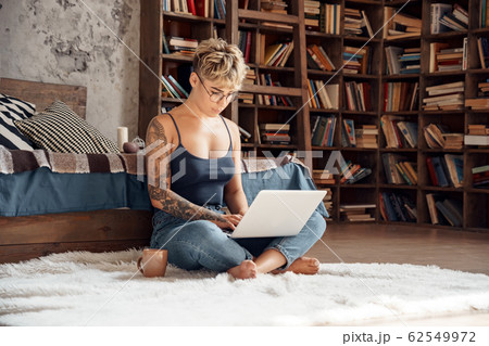 Side Hustle. Young woman short hair in glasses sitting on floor working online on laptop concentrated with cup of coffee Side Hustle. Young woman short hair in glasses sitting on floor working online on laptop concentrated with cup of coffee 62549972
