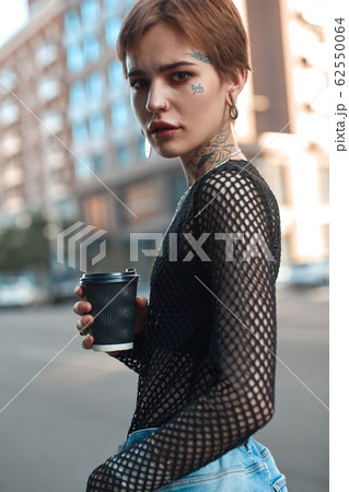 Urban Style. Young stylish woman standing outdoors with cup of coffee looking camera curious close-up Urban Style. Young stylish woman standing outdoors with cup of coffee looking camera curious close-up 62550064