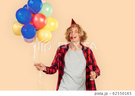 Freestyle. Teen blond boy in birthday cap standing isolated on nude color with balloons blowing party horn closed eyes laughing cheerful 62550165