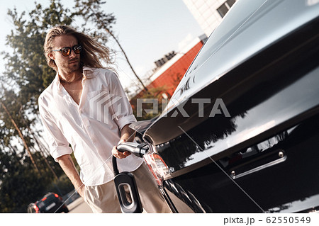 Transportation. Young man with long hair in sunglasses traveling by electric car stop at chraging station holding charger watching concerned 62550549