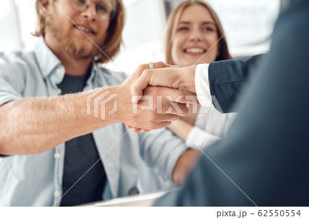Car Salon. Young couple sitting at table man shaking hands with agent buying car close-up smiling happy Car Salon. Young couple sitting at table man shaking hands with agent buying car close-up smiling happy 62550554