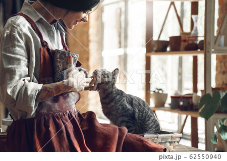 Craftsperson Concept. Young woman making pottery indoors standing with cup scratching cat smiling happy little friend 62550940