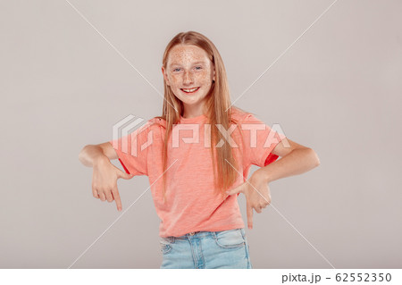 Inclusive Beauty. Girl with freckles standing isolated on grey pointing down at copy space smiling cheerful Inclusive Beauty. Girl with freckles standing isolated on grey pointing down at copy space smiling cheerful 62552350