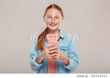 Inclusive Beauty. Girl with freckles in denim jacket standing isolated on grey with smartphone looking camera excited Inclusive Beauty. Girl with freckles in denim jacket standing isolated on grey with smartphone looking camera excited 62552351