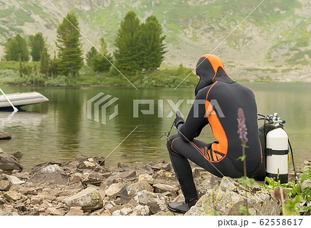a guy in a diver suit is sitting on rocks on the lake shore 62558617