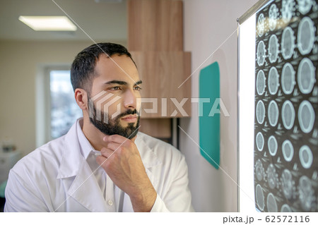 Bearded young doctor in a white robe analyzing MRI results Bearded young doctor in a white robe analyzing MRI results 62572116