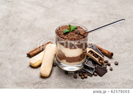 Classic tiramisu dessert in a glass cup, pieces of chocolate and savoiardi cookies on concrete background Classic tiramisu dessert in a glass cup, pieces of chocolate and savoiardi cookies on concrete background 62579203
