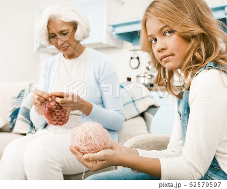 Granddaughter Helping Her Grandmother To Knit While They Sitting On White Sofa Granddaughter Helping Her Grandmother To Knit While They Sitting On White Sofa 62579597