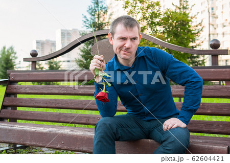A young man sitting on a bench with a rose in his hands looks with contempt at passers-by 62604421