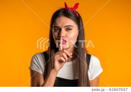 Smiling woman with long hair holding finger on her lips over yellow background. Gesture of shhh, secret, silence. Close up. 62607687
