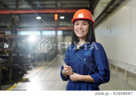 Pretty young smiling engineer in uniform and protective helmet holding tablet Pretty young smiling engineer in uniform and protective helmet holding tablet 62608687