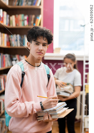 Teenager with backpack, stack of books and pencil standing in college library 62609062