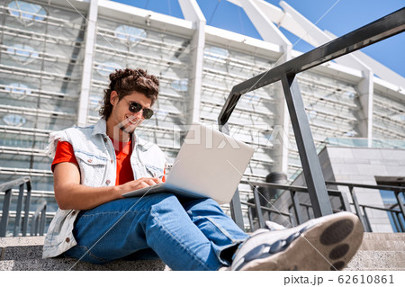 Freestyle. Guy in sleeveless jean jaket and sunglasses outdoors sitting on stairs browsing internet on laptop smiling happy bottom view 62610861