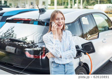 Transportation. Young woman on electric car having stop at charging station leaning on vehicle talking on smartphone laughing cheerful while charging 62610891