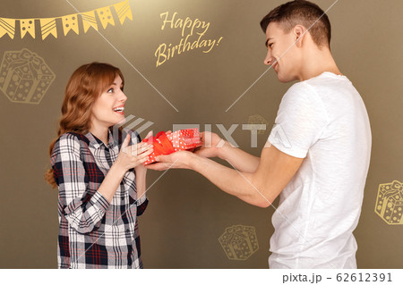 Celebration Concept. Young boyfriend giving gift to touched girlfriend standing isolated on grey close-up Celebration Concept. Young boyfriend giving gift to touched girlfriend standing isolated on grey close-up 62612391