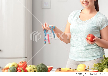 Cooking Meal. Chubby girl standing in kitchen with tape measure and apple posing to camera smiling cheerful 62613453