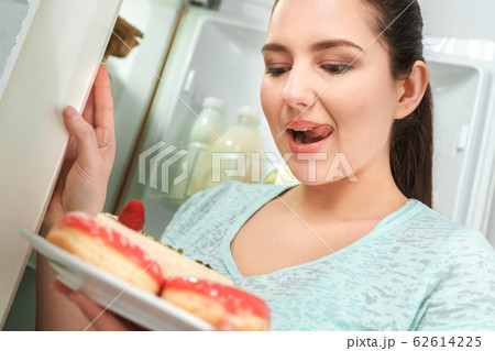 Body Care. Chubby girl standing in kitchen near fridge looking at plate with doughnuts and cheesecake smiling excited close-up Body Care. Chubby girl standing in kitchen near fridge looking at plate with doughnuts and cheesecake smiling excited close-up 62614225