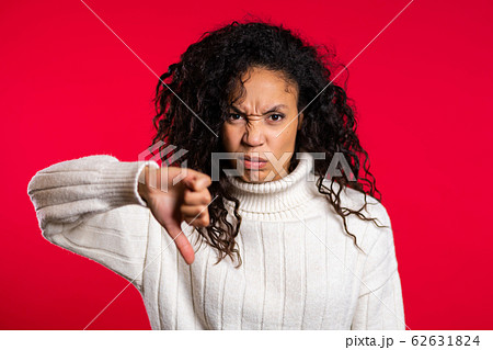 Young pretty mixed race woman standing on red studio background expressing discontent and showing thumb down gesture at camera. Portrait of girl with sign of dislike 62631824
