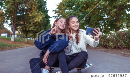 Girls girlfriends teenagers, summer park, photos smartphone, happy smiling, having fun playing. Selfie phone. Record video message, online social networks Internet. Skateboard, trees road background. Girls girlfriends teenagers, summer park, photos smartphone, happy smiling, having fun playing. Selfie phone. Record video message, online social networks Internet. Skateboard, trees road background. 62635987