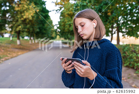 Serious teenage girl schoolgirl, summer park, holding smartphone in hand, watching video headphones. Rest in park after school college. Emotions understanding learning, video lesson. 62635992