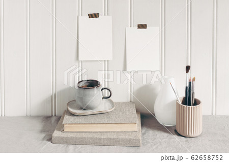 Blank paper cards taped on white wooden wall. Stationery mockup scene with paint brushes, pencils in ceramic holder, cup of coffee and books on linen tablecloth. Artistic scene. Creative background Blank paper cards taped on white wooden wall. Stationery mockup scene with paint brushes, pencils in ceramic holder, cup of coffee and books on linen tablecloth. Artistic scene. Creative background 62658752