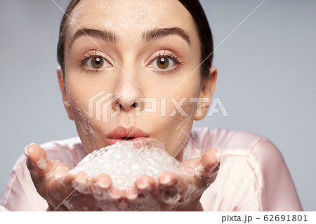 Beautiful young woman blowing foam while washing face Beautiful young woman blowing foam while washing face 62691801