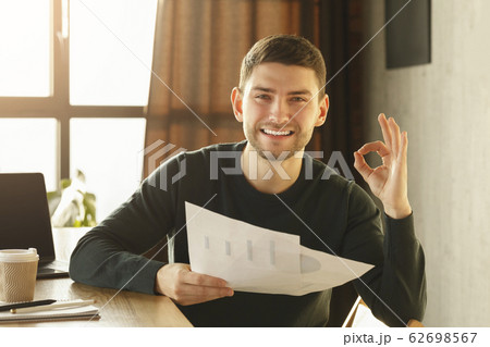 Guy Gesturing Okay Holding Papers With Charts Sitting At Workplace Guy Gesturing Okay Holding Papers With Charts Sitting At Workplace 62698567