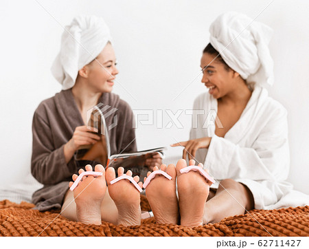 Girlfriends in bathrobes making pedicure at home, reading magazine 62711427