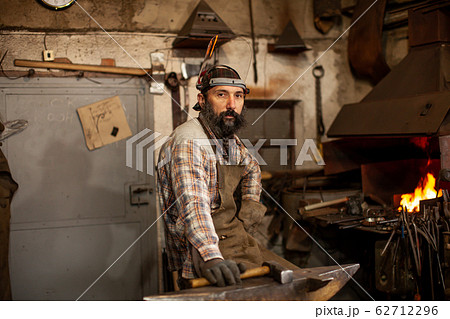 The portrait of bearded blacksmith preparing to work in his workshop. Small business. The portrait of bearded blacksmith preparing to work in his workshop. Small business. 62712296