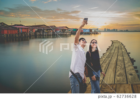 Happy couple taking selfie with cell mobile phone in Chew Jetty fisherman village at sunset. Happy couple taking selfie with cell mobile phone in Chew Jetty fisherman village at sunset. 62736628