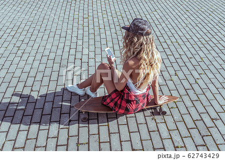 Beautiful girl with long hair holding mobile phone in his hand, in summer in city casual clothes, swimsuit, baseball cap shirt. Long board, skate, social networks online application. Beautiful girl with long hair holding mobile phone in his hand, in summer in city casual clothes, swimsuit, baseball cap shirt. Long board, skate, social networks online application. 62743429